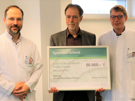 Prof. Thomas H. Ittel, Vorstandsvorsitzender der Uniklinik und der Stiftung Universitätsmedizin Aachen (Mitte), überreicht den Scheck an Priv.-Doz. Dr. med. Daniel Delev (links) und Univ.-Prof. Dr. med. Hans Clusmann, Direktor der Klinik für Neurochirurgie.