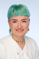 Portraitfoto einer Person mit türkisen Haaren in weißer medizinischer Kleidung, die freundlich in die Kamera lächelt.
