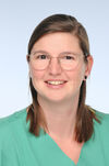 Annika Bell Porträtbild von Annika Bell einer Frau mit schulterlangem braunem Haar, die eine grüne medizinische Berufskleidung trägt. Sie hat ein freundliches Lächeln, trägt eine runde Brille mit dünnem Metallrahmen und schwarze Ohrstecker. Der Hintergrund ist hellblau