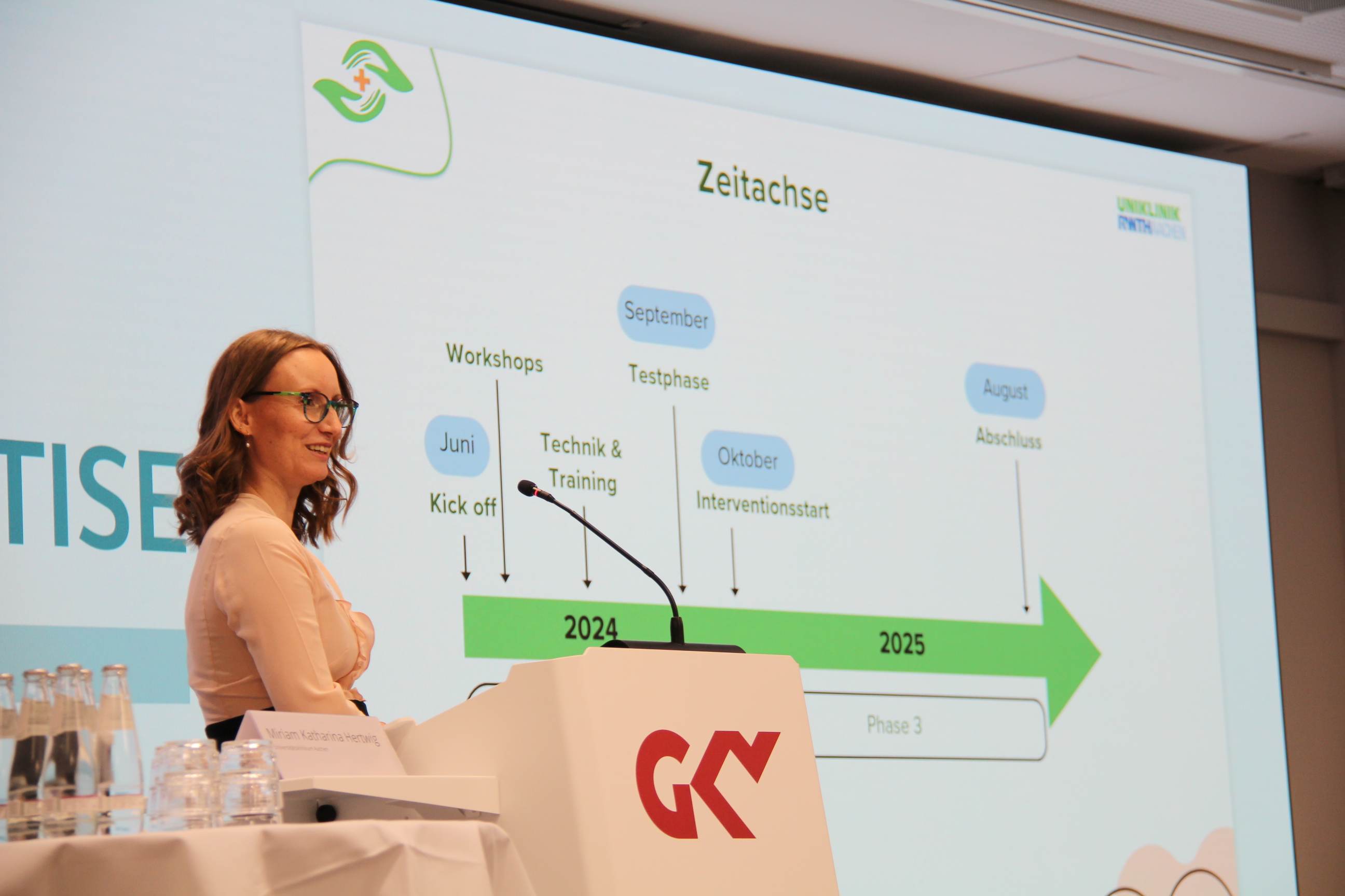 Ein Frau mit braunen Haaren und Brille steht vor einer Leinwand, auf der eine Grafik mit einem Pfeil dargestellt ist. 