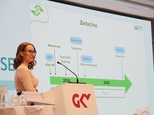 Ein Frau mit braunen Haaren und Brille steht vor einer Leinwand, auf der eine Grafik mit einem Pfeil dargestellt ist. 