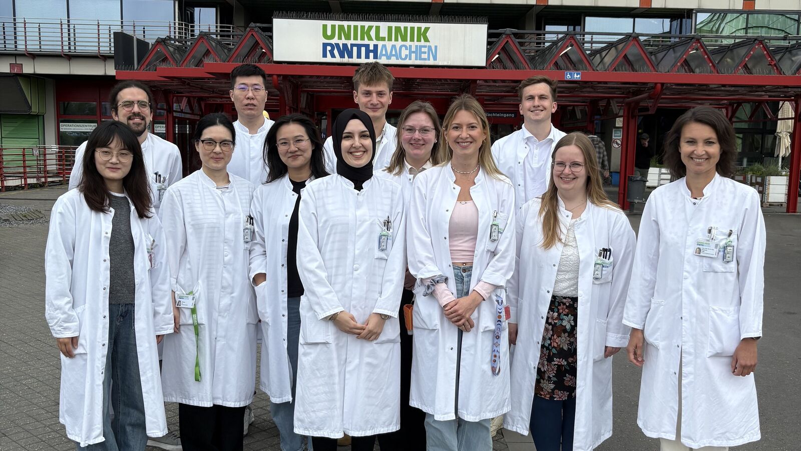 Teamfoto von mehreren Frauen und Männern in weißen Laborkitteln draußen vor dem Haupteingang der Uniklink RWTH Aachen.
