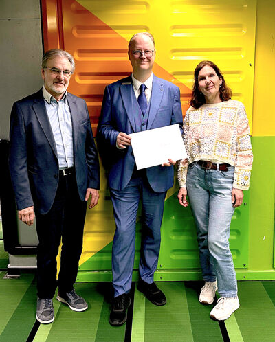 Gruppenfoto von drei Personen. Eine Frau und zwei Männer posieren für die Kamera mit einem symbolischen Scheck