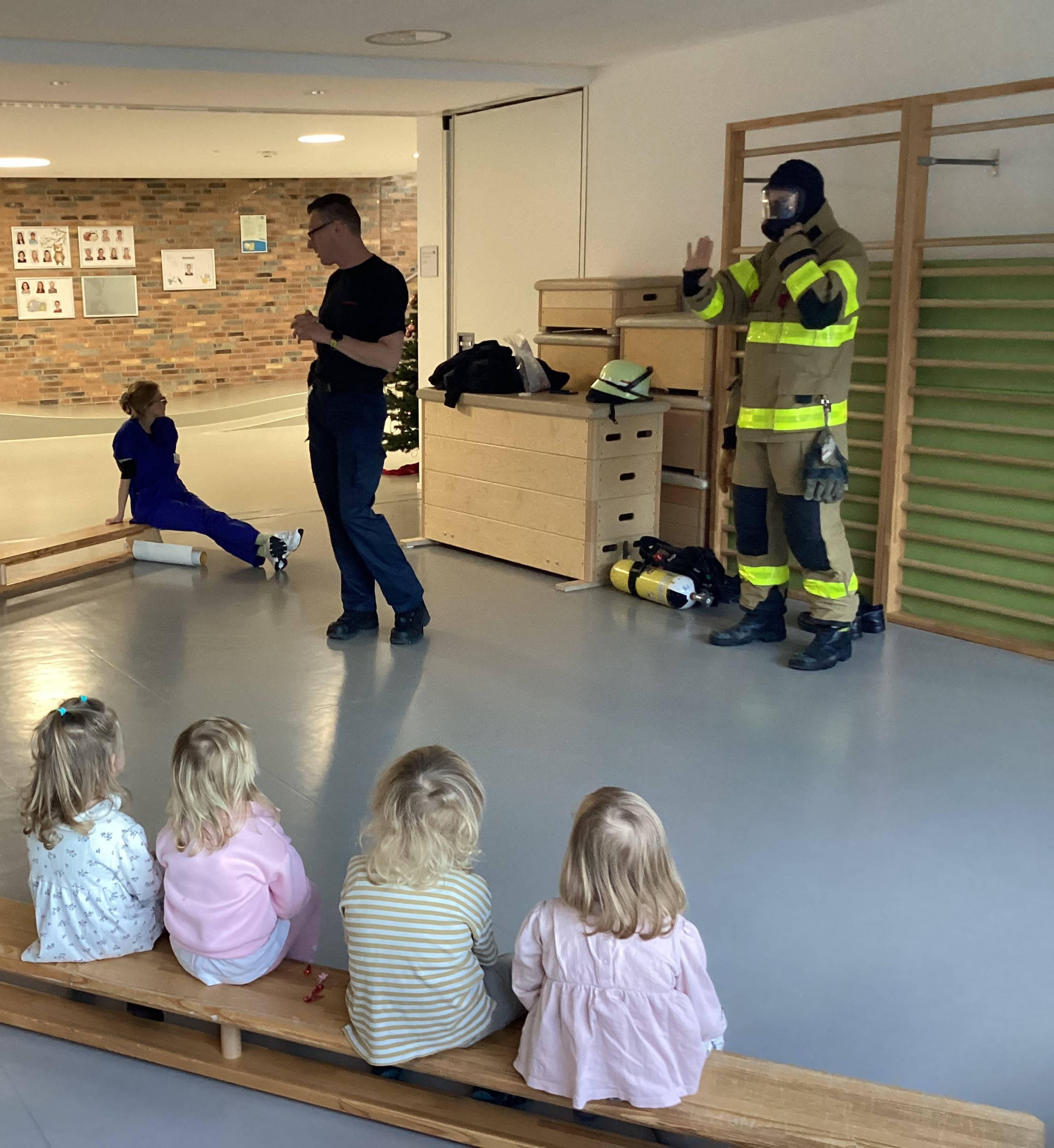 Kinder sitzen auf einer Holzbank und hören den Erklärungen eines Feuerwehrmanns zu