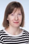 Aleksandra Boiko Porträtaufnahme einer Person mit kurzem braunem Haar und schwarz-weiß gestreiftem Oberteil vor hellblauem Hintergrund
