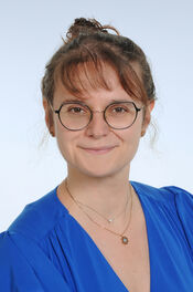 Porträtfoto einer Frau mit braunem Haar, Zopf und Brille, die ein blaues Oberteil trägt und freundlich in die Kamera lächelt.