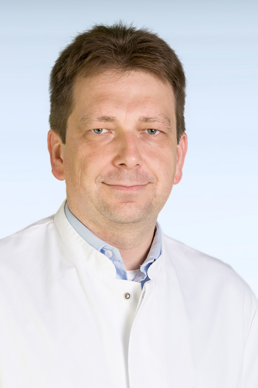 Porträtfoto eines Mannes mit kurzen braunen Haaren in einem weißen Arztkittel, der freundlich in die Kamera blickt.