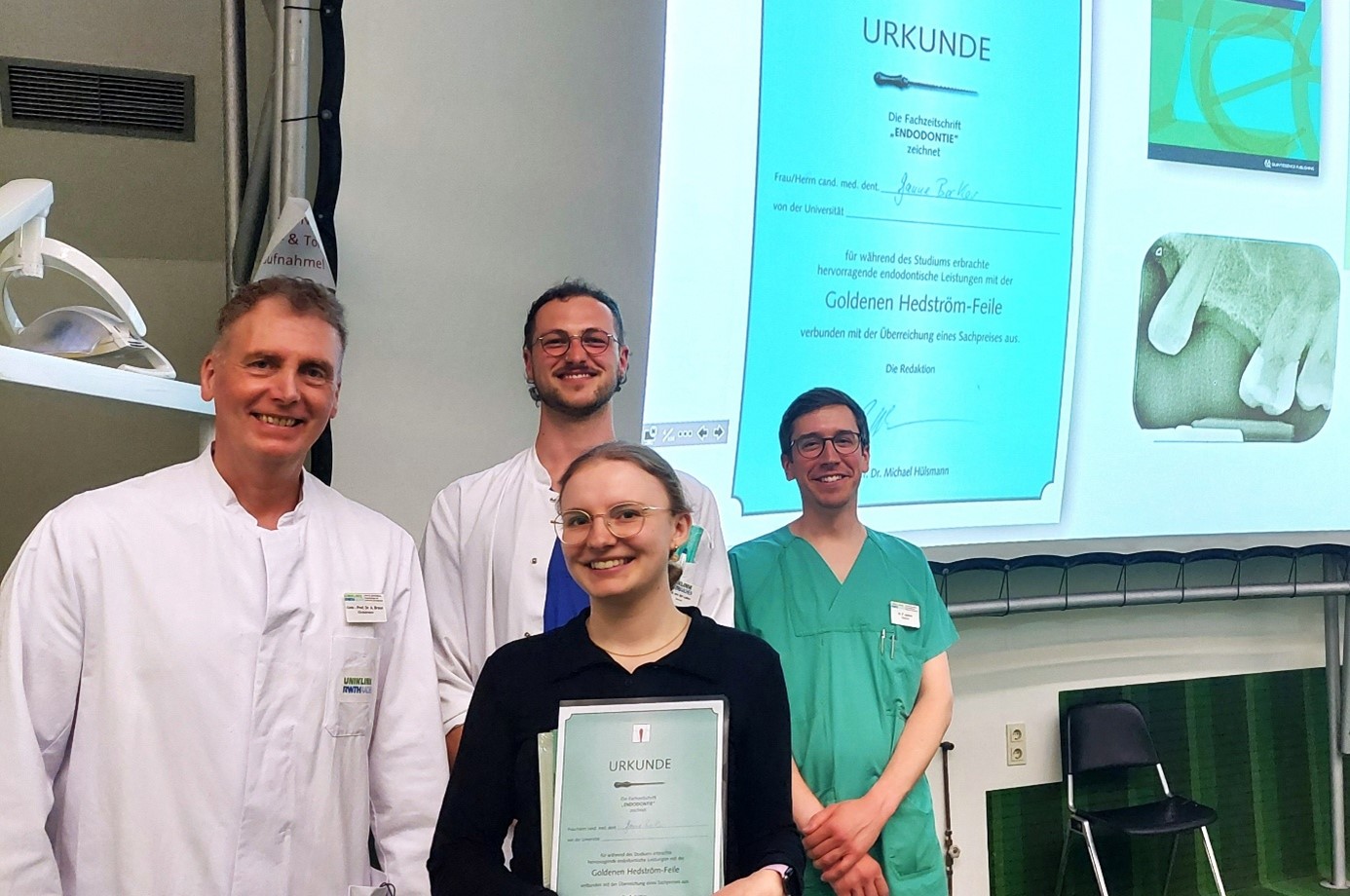 Vier Personen in medizinischer Kleidung stehen lächelnd in einem Hörsaal. Eine junge Frau in der Mitte hält eine Urkunde in den Händen – sie wurde für besondere Leistungen im Bereich der Endodontie mit der "Goldenen Hedström-Feile" ausgezeichnet. Im Hintergrund ist eine Projektion der Urkunde sowie ein Röntgenbild eines Zahns zu sehen.