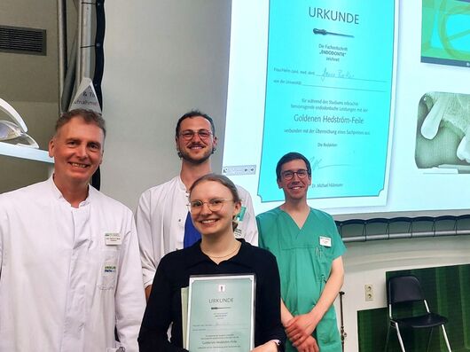 Vier Personen in medizinischer Kleidung stehen lächelnd in einem Hörsaal. Eine junge Frau in der Mitte hält eine Urkunde in den Händen – sie wurde für besondere Leistungen im Bereich der Endodontie mit der "Goldenen Hedström-Feile" ausgezeichnet. Im Hintergrund ist eine Projektion der Urkunde sowie ein Röntgenbild eines Zahns zu sehen.