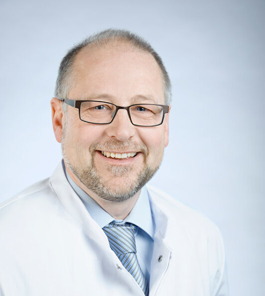 apl. Prof. Dr. rer. nat. Albrecht Eisert Porträtfoto eines Mannes mit hellen Haaren, Bart und Brille, der einen weißen Kittel trägt und freundlich in die Kamera lächelt.