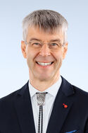 Portraitfoto eines Mannes mit kurzen grauen Haaren mit einer Bolo-Krawatte in einem dunklen Anzug, der freundlich in die Kamera lächelt.